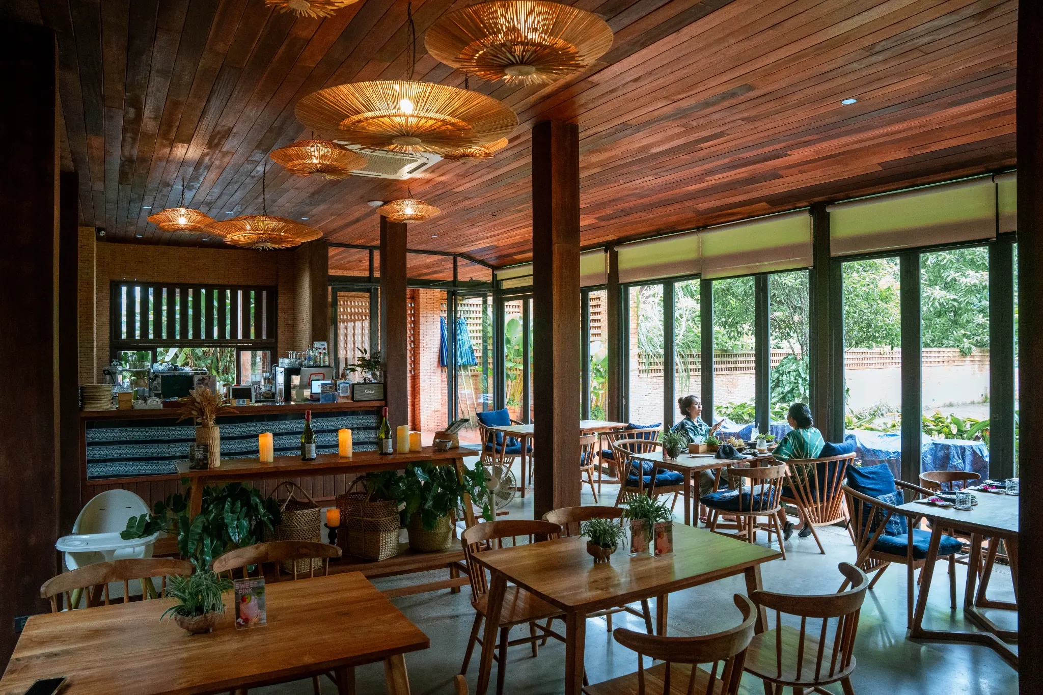 Warm wooden dining room. Wooden tables and chairs sit beneath woven pendant lights, beside large windows and a service counter.